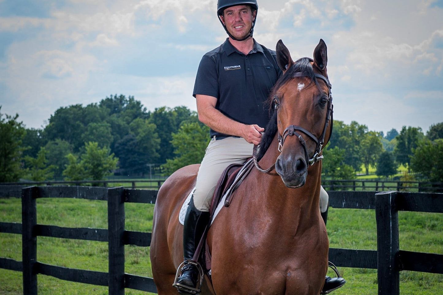 Man in riding gear sits atop Carlchen W in a grassy paddock, smiling at the camera with trees and a wooden fence.