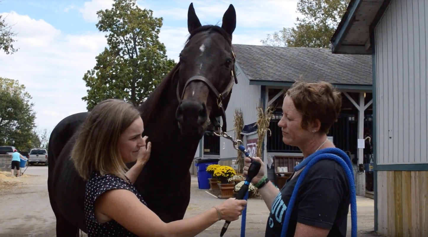 Two women stand on either side of a dark horse during an outdoor interview with barns and trees in the background.