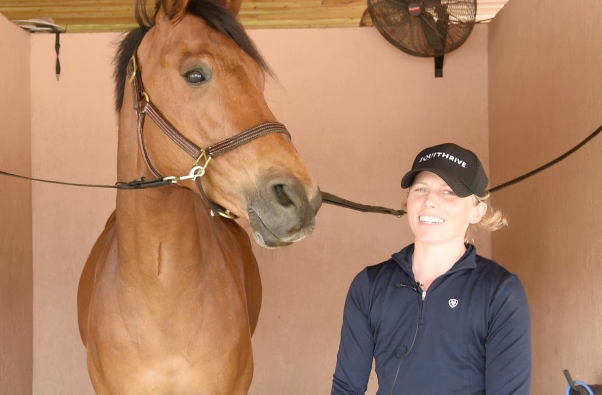 Emily smiling in an Equithrive cap stands beside a Audi in a stable with fans and light pink walls in the background.