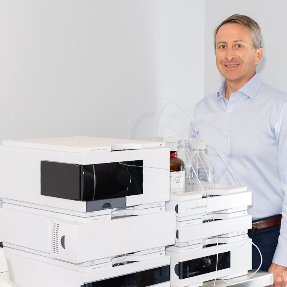 Dr. Lawless in a light blue shirt stands next to a laboratory instrument with multiple stacked white modules and tubing.