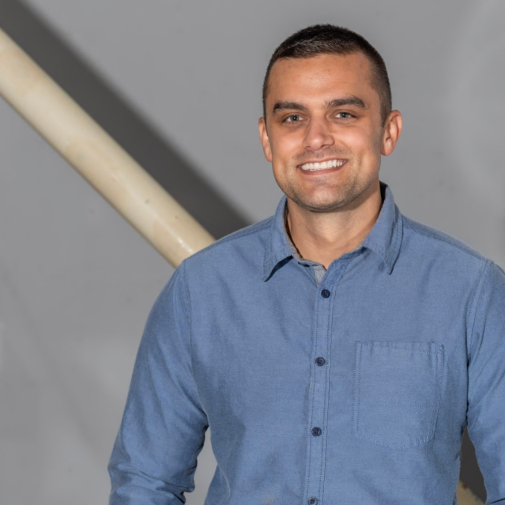 Joshua Hager wearing a blue button-up shirt stands in front of a neutral gray background with a diagonal railing behind him.