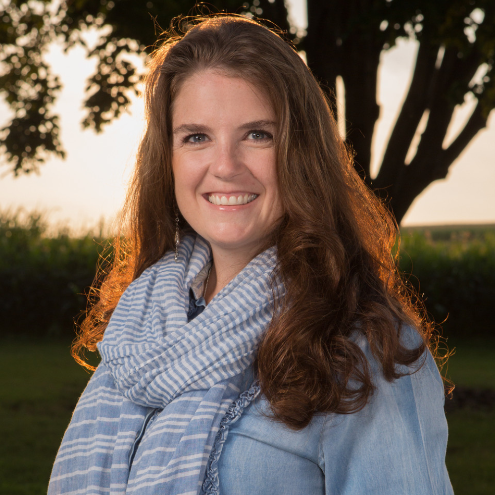 Molly Wagner with long brown hair wearing a blue denim shirt and striped scarf stands outdoors with trees and a sunset.